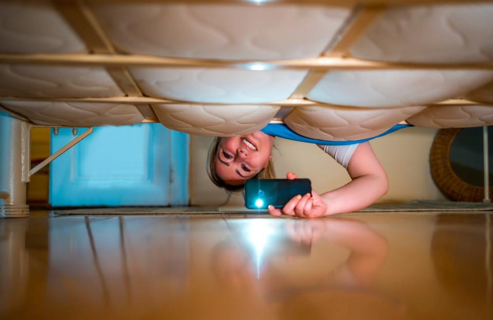 Young smiling woman looking under the bed, using flashlight in search