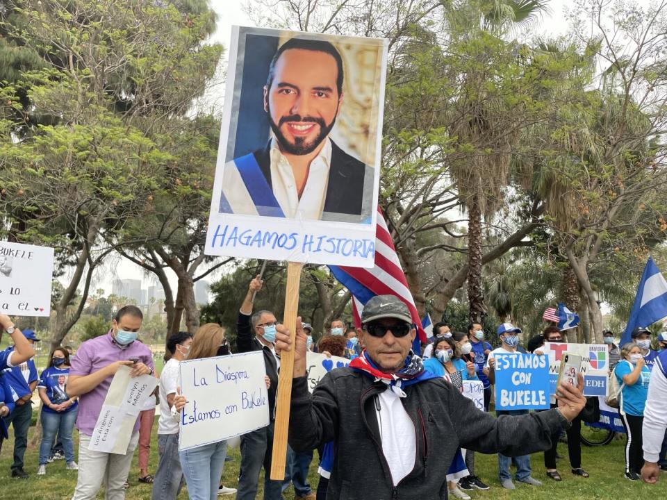 A man in dark clothes, standing in a crowd, holds a placard with an image of a smiling bearded man in a dark suit