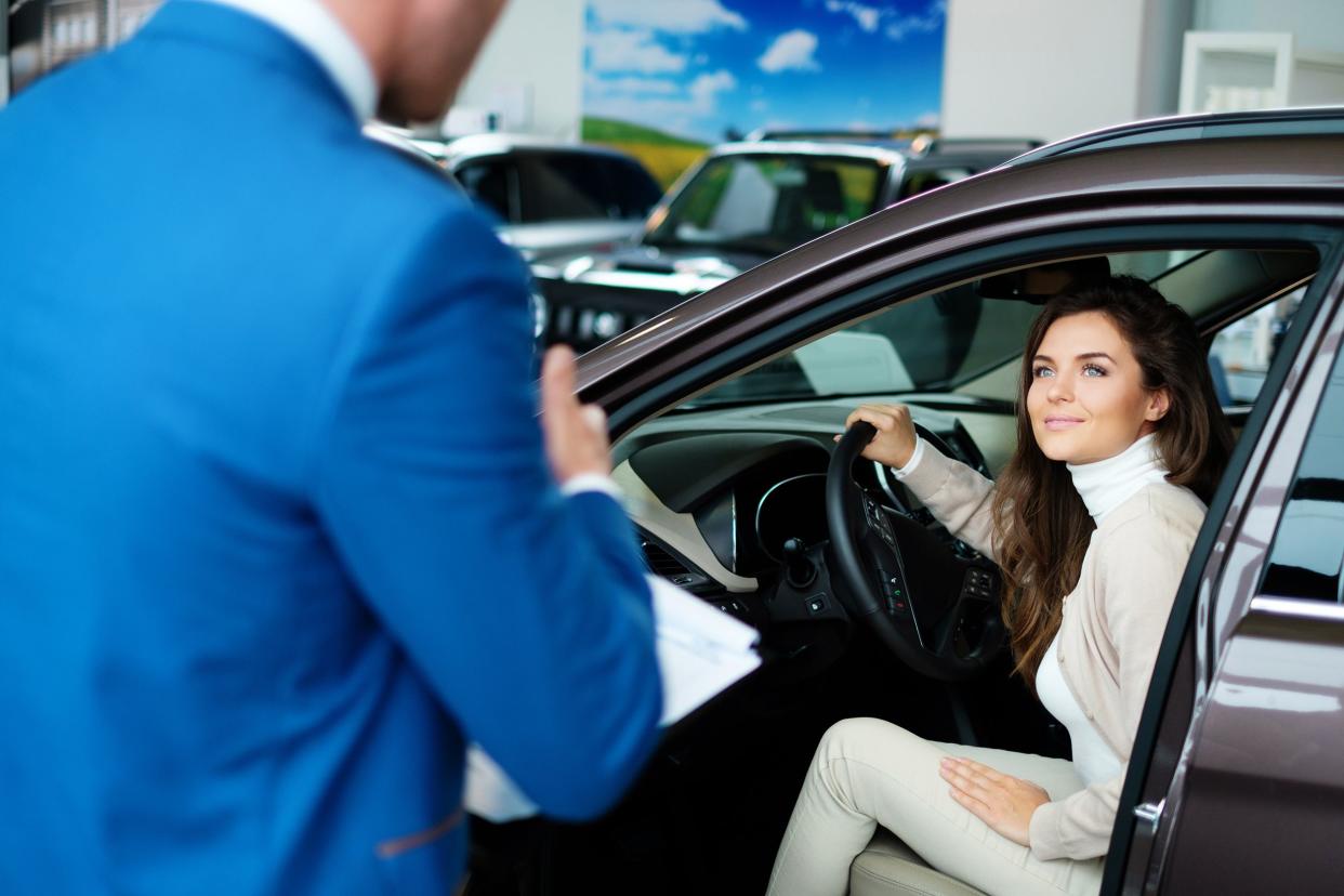 Woman is in car in car dealership showroom looking at male car dealer as he gives her information about the vehicle with other vehicles in the background