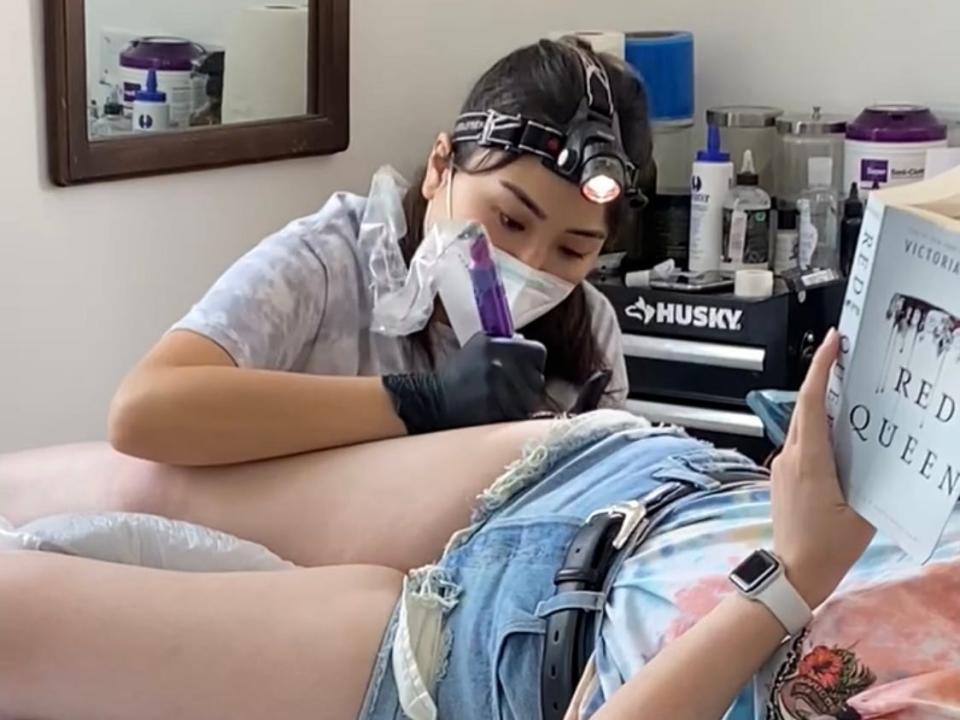 The writer tattooing a client on their leg while the client reads a book