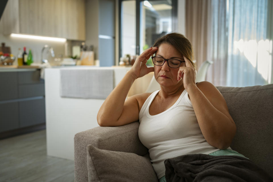 A woman in a white tank top and glasses sits on a sofa, holding her temples with a pained expression in a modern, sunlit kitchen and living room