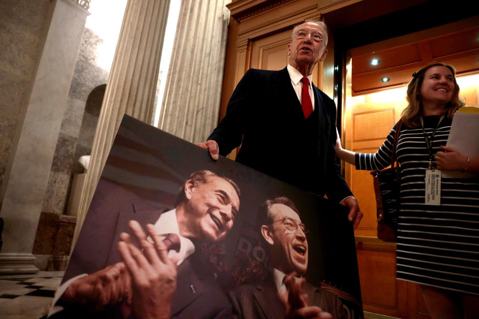 Republican Sen. Chuck Grassley of Iowa totes a poster-sized picture of himself and late-Republican Sen. Bob Dole of Kansas while standing outside the Senate chamber as scheduler Jennifer Heins shoots reporters a smile in December 2021.