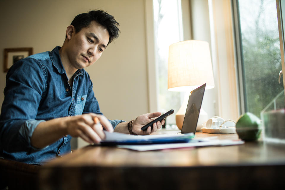 A man (early 30s) working in his home office, investing concept. He's looking at papers while on his computer