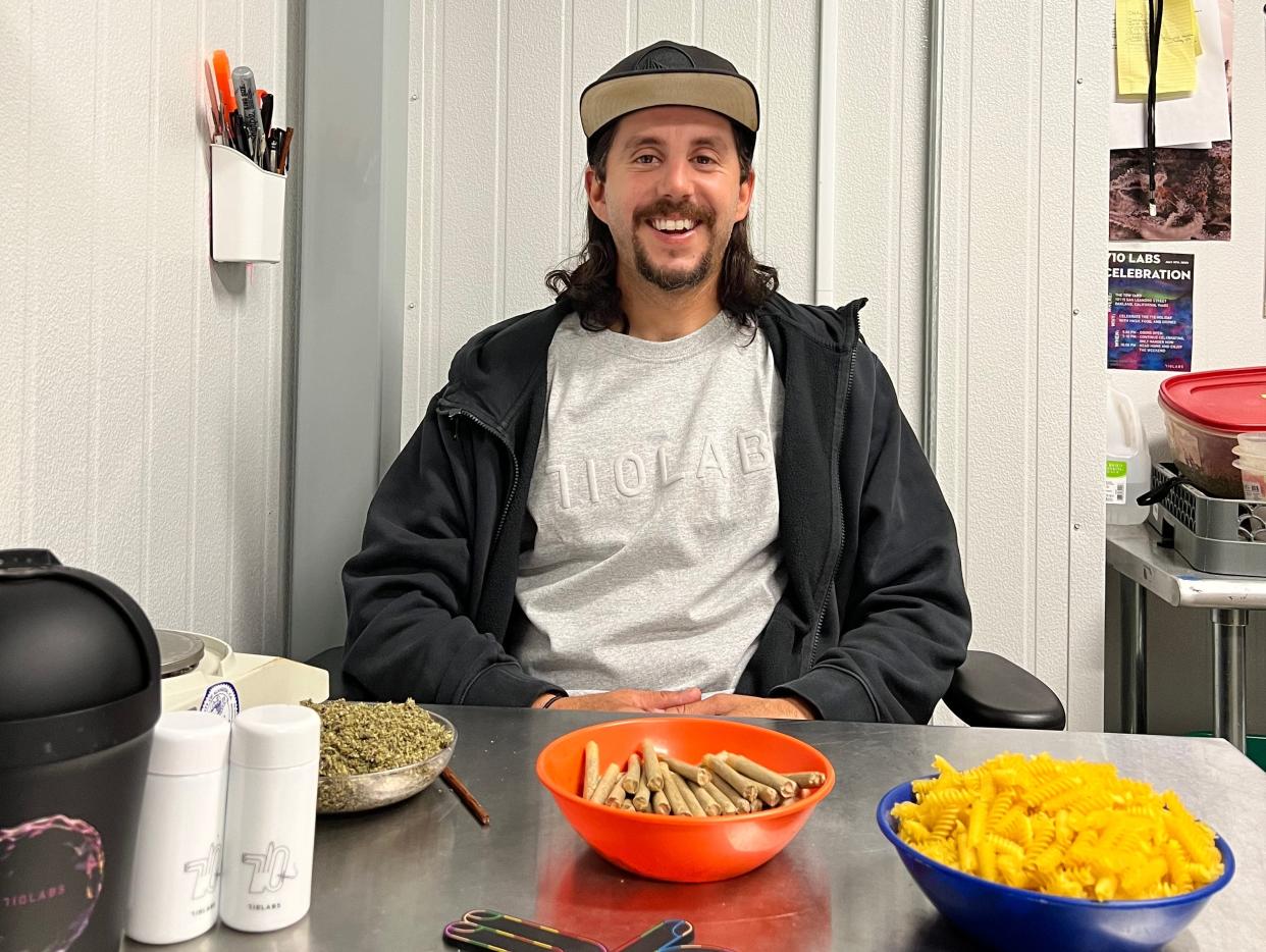 Photograph of Thomas Indigaro smiling sat behind a table with bowls of rolled joints and rotini pasta filters.