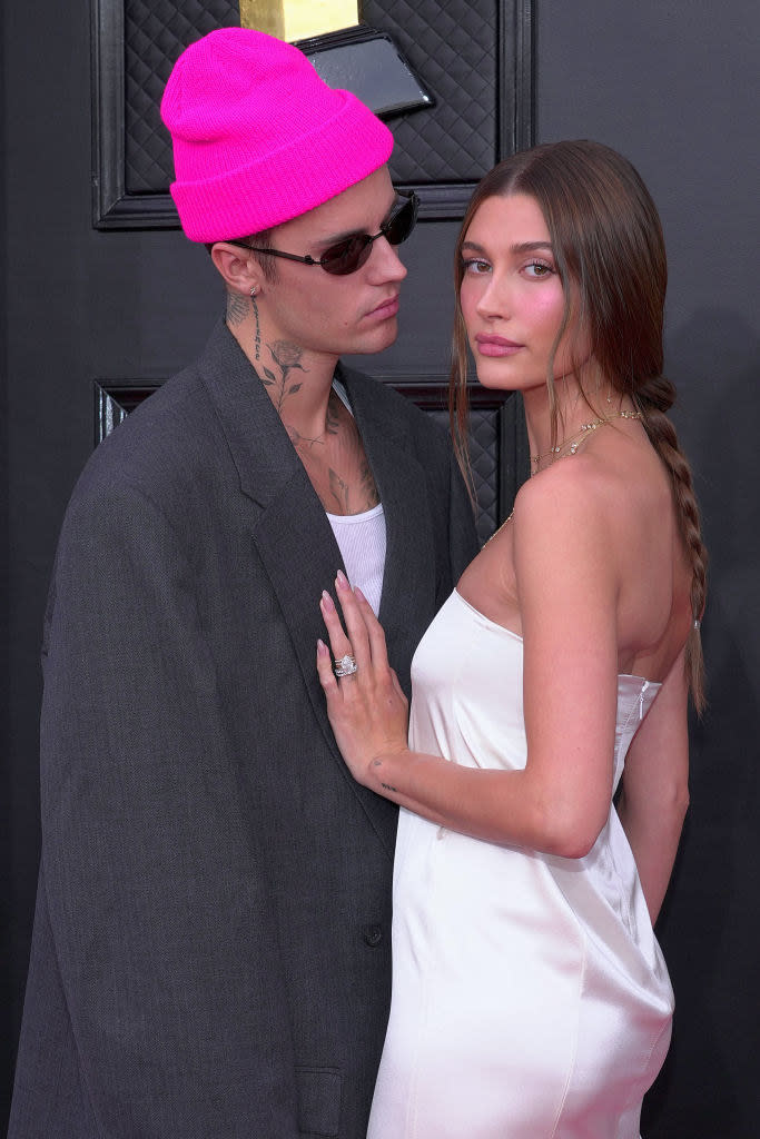 Justin, wearing a beanie and sunglasses, looking at Hailey, who's wearing a slinky, strapless gown and has her hand on his chest