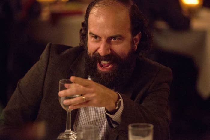 Man with beard in suit laughs holding a glass at a table in a scene from a TV show