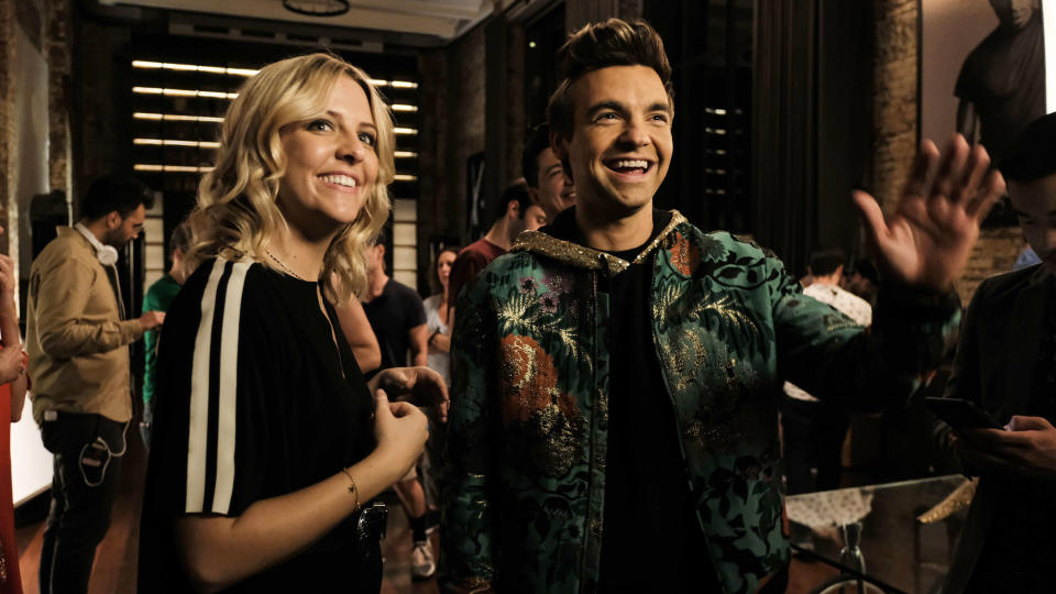 Heidi Gardner and Andrew Rannells in a lively gathering, smiling and engaging with others in the background. Andrew wears a patterned jacket