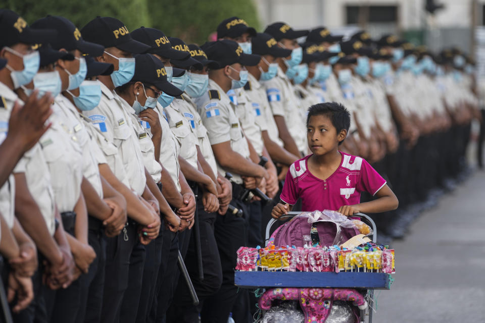 A teenager selling candy walks past police standing guard outside the National Palace where people rally in support of anti-corruption Prosecutor Juan Francisco Sandoval in Guatemala City, Saturday, July 24, 2021. Sandoval fled Guatemala late Friday, arriving in neighboring El Salvador just hours after he was removed from his post. (AP Photo/Moises Castillo)