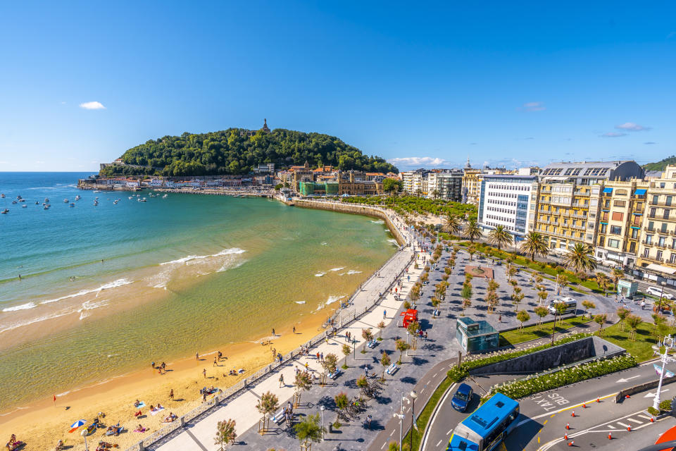 Aerial view of a vibrant, populated beach beside a busy promenade and a scenic coastal city with lush hills in the background