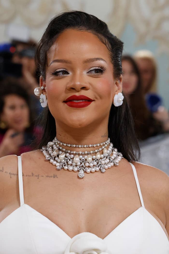 Rihanna smiles wearing a layered pearl necklace and white dress with floral details at a formal event. She has pearl earrings that match her necklace