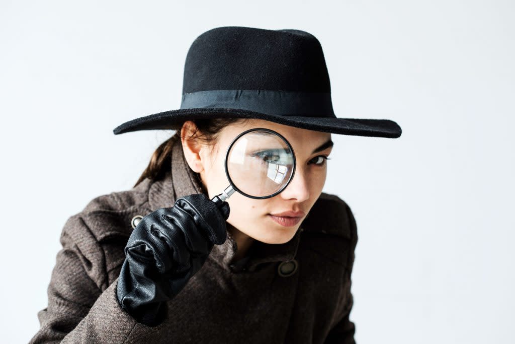 A woman looks through a magnifying glass while wearing a coat, black gloves and a big hat.