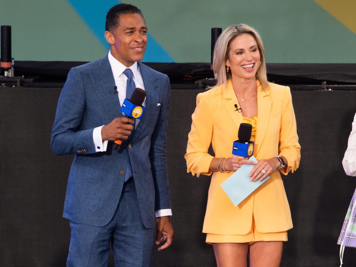 T.J. Holmes, in a blue suit, smiles while standing next to fellow "GMA" host Amy Robach, in a yellow blazer and skirt, on stage.