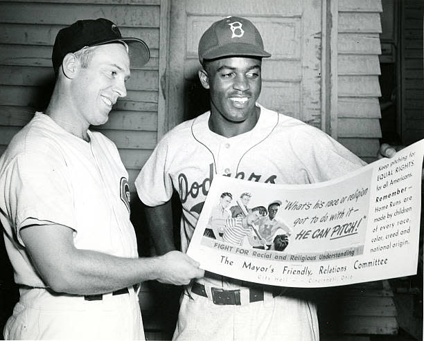 American baseball players Danny Litwhiler (left) of the Cincinnati Reds and Jackie Robinson (1919 - 1972) of the Brooklyn Dodgers pose together as they smile and hold a poster form the 'Mayor's Friendly Relations Committee,' Cincinnati, Ohio, May 11, 1984. The poster features an illustration of a group of boys and the text 'What's his race or religion go to do with it--he can pitch!', 'Fight for Racial and Religious Understanding', and 'Keep pitching for EQUAL RIGHTS for all Americans. Remember--Home Runs are made by children of every race, color, creed and national origin.' (Photo by Betz-Marsh Studio/Cincinnati Museum Center/Getty Images)