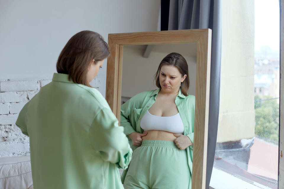 Vertical shot, size plus woman in casual clothes or pyjamas staring in full length mirror at home, looking, check body condition feels frustrated and dissatisfied by overweight, touch fat accumulations frowning. Diet oneself concept