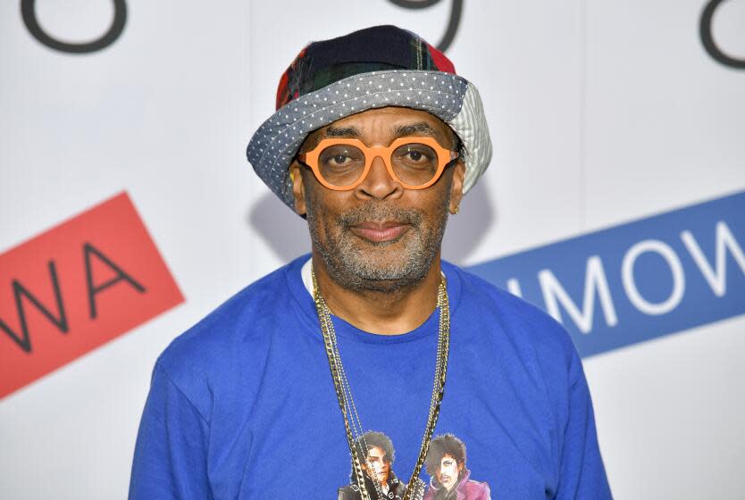 Spike Lee in thick orange glasses, a patterned bucket hat, bright blue shirt and a long gold chain looking straight ahead