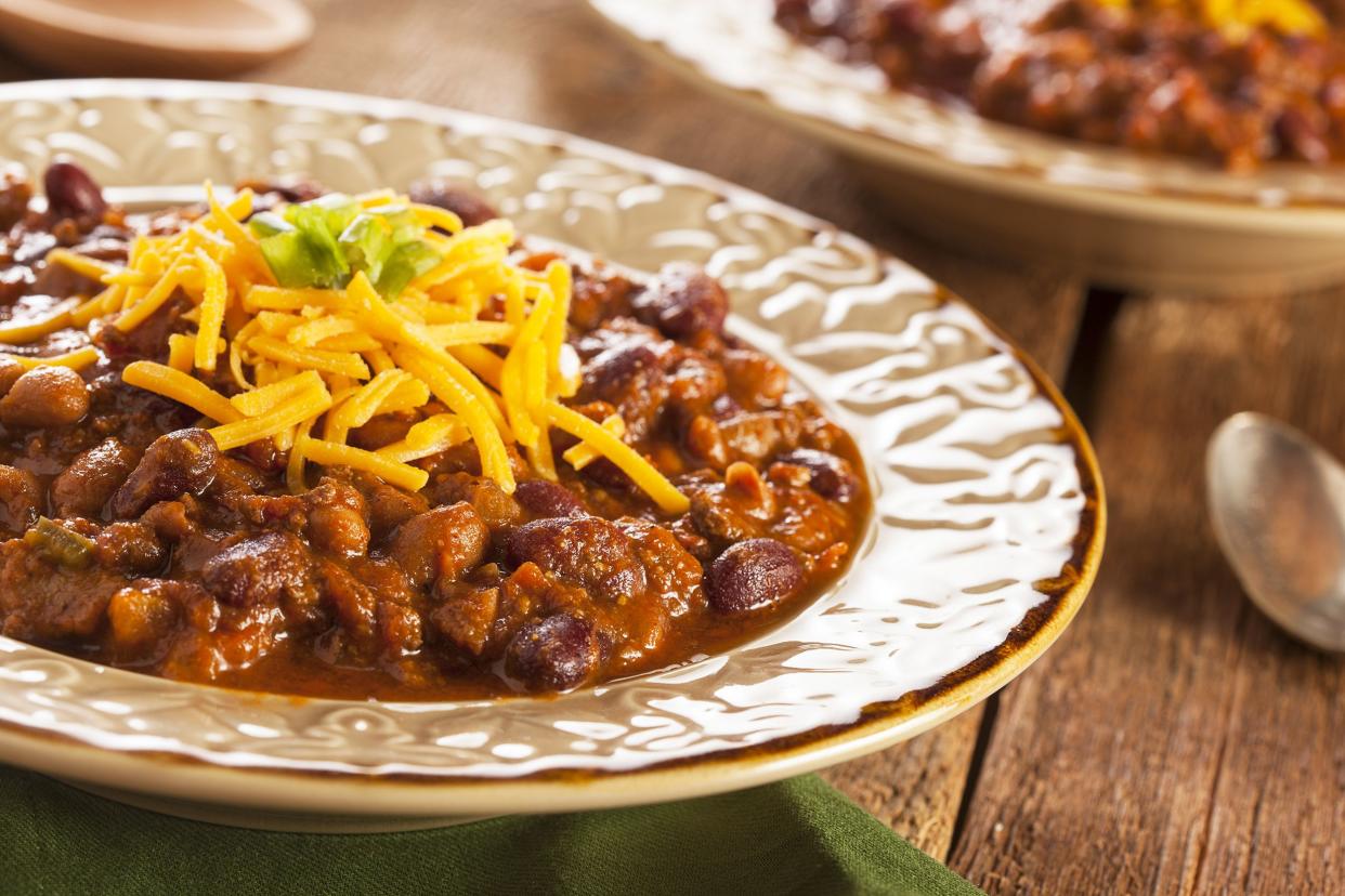 Focus on ground beef chili in a wide-rimmed beige ceramic bowl, on an olive napkin on a rustic wooden table with another bowl of beef chili blurred in the background