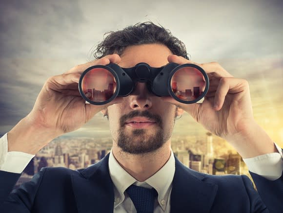 Man in suit and tie looking through binoculars with a cityscape in the background