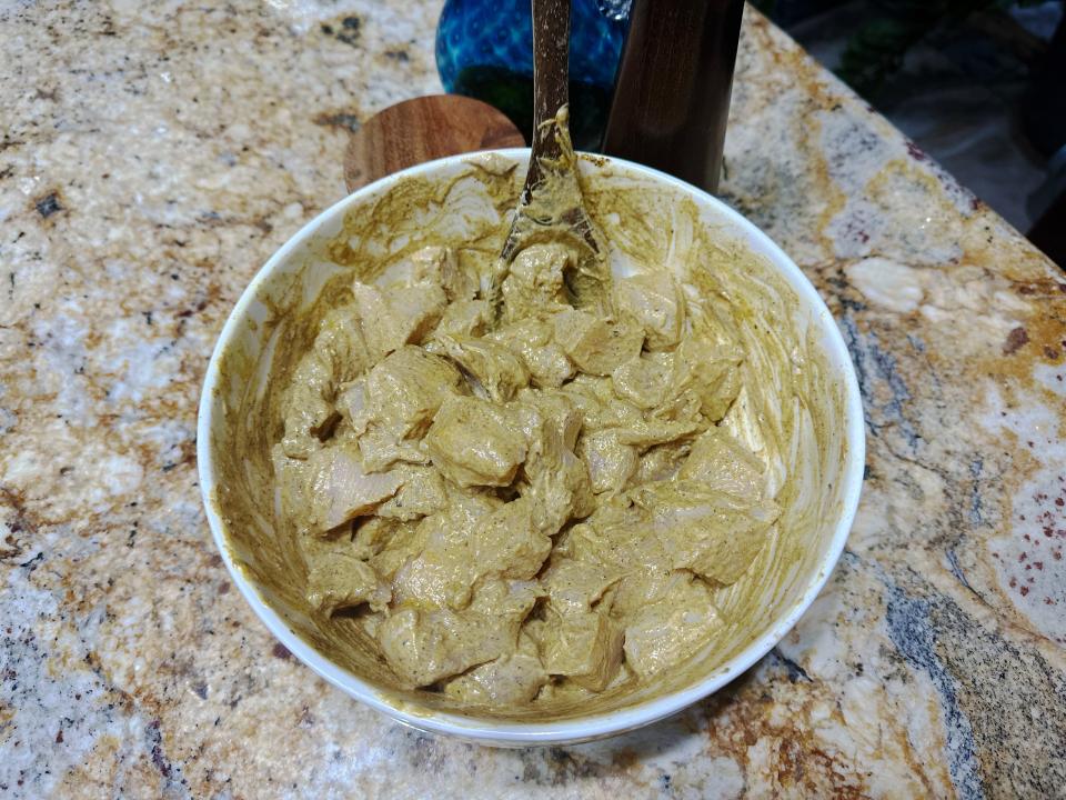 Cubed, raw chicken covered with a yellow-brown marinade in a white bowl with a spoon sticking out.