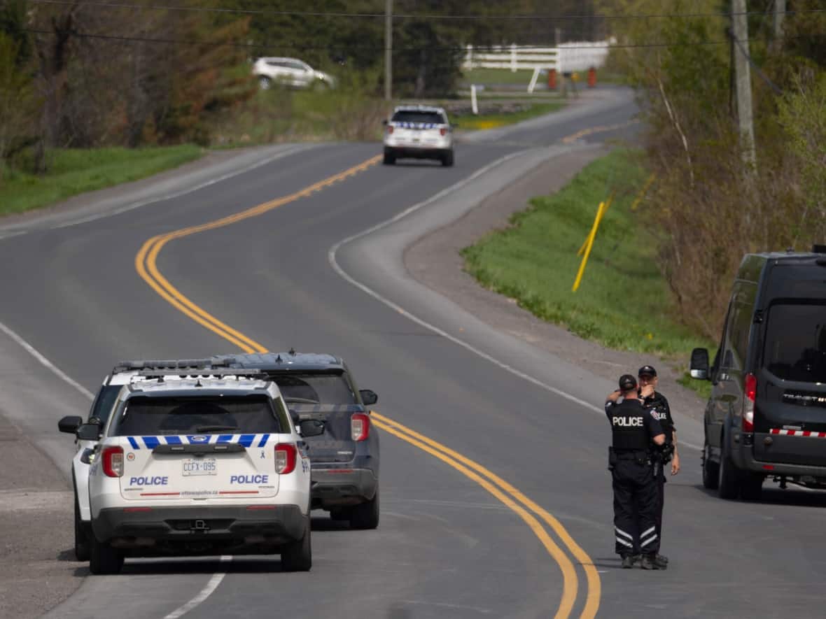 Police officers stand on a rural highway on May 11, 2023, near Bourget, Ont., where three Ontario Provincial Police officers were shot earlier in the day. One of those officers, Sgt. Eric Mueller, later died in hospital. (Jean Delisle/CBC - image credit)
