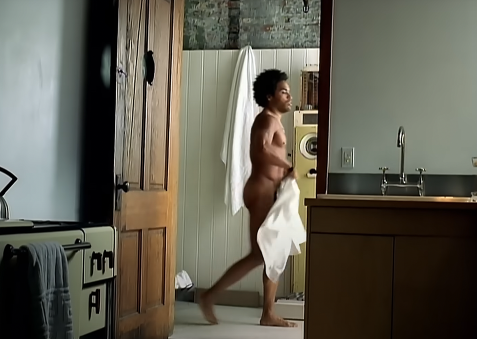 Naked person holding a towel seen walking in a rustic, modern bathroom setup. The person's identity is not recognizable
