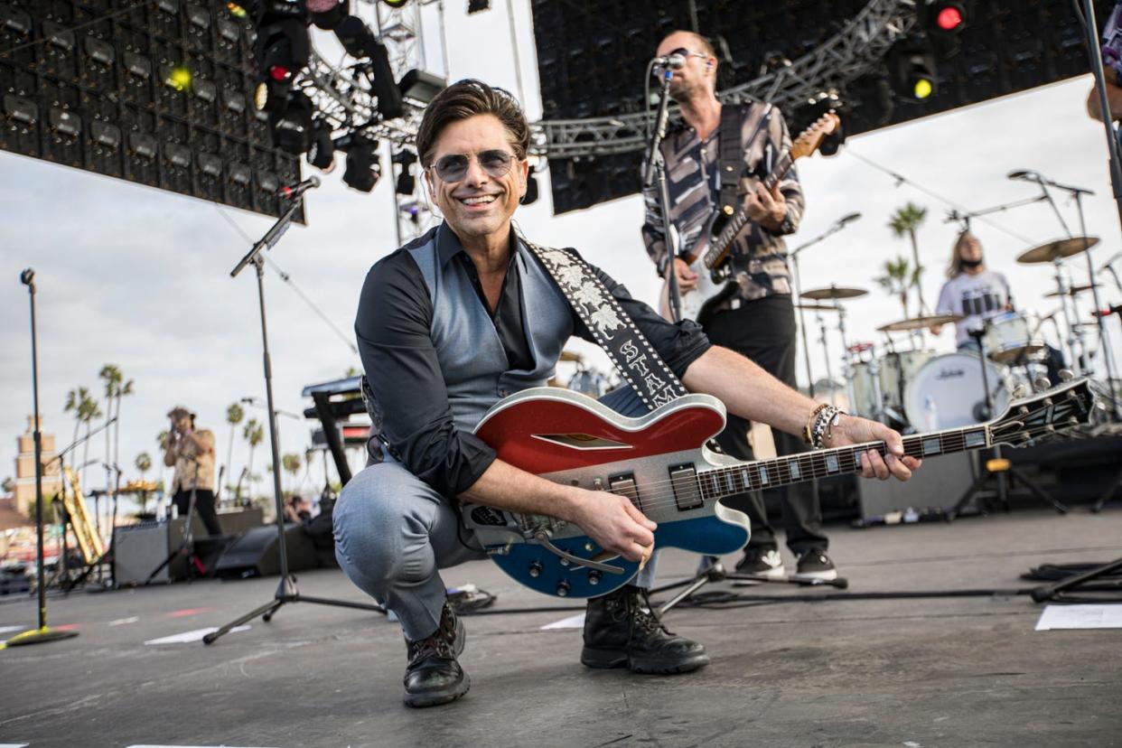 john stamos smiles while playing an electric car on stage, he wears a gray vest and pants, a black long sleeve shirt, and sunglasses, musicians play behind him