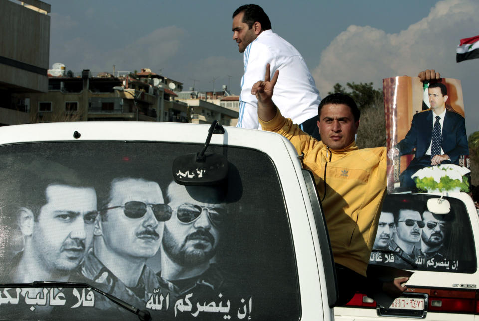 FILE - In this March 25, 2011, file photo, pro-Syrian President Bashar Assad protester, holds Bashar Assad's poster as he sits outside a mini van window during a pro-Assad demonstration, in Damascus, Syria. More than 130,000 people have died in three years, and more than a quarter of the country's population now live as refugees, either displaced internally or in neighboring countries. An untold number of people have been subjected to the trauma of losing loved ones or witnessing lives blown apart. Posters on the back window are for Syrian President Bashar Assad, center, and his two brothers Maher Assad, left, and Bassel Assad, right, with Arabic reads, "If God triumphs you, no one can defeat you." (AP Photo/Hussein Malla, File)