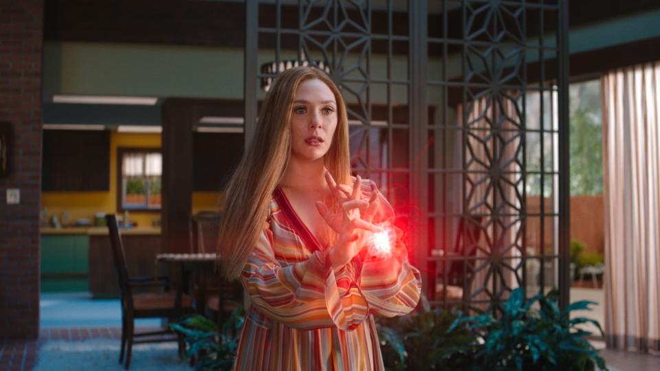 Elizabeth Olsen, in a striped '70s-era dress on a period set, holds her hands in front of her as fire forms between them.