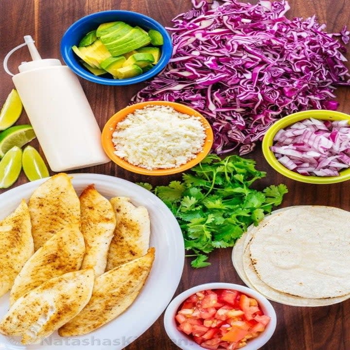 Taco ingredients on a tabletop including cabbage, lime, tortillas, salsa, onion, and fish fillets.