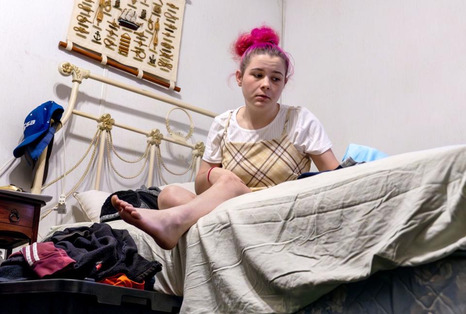 Framed from a low angle, a teen girl sits on a bed, staring off with a forlorn look