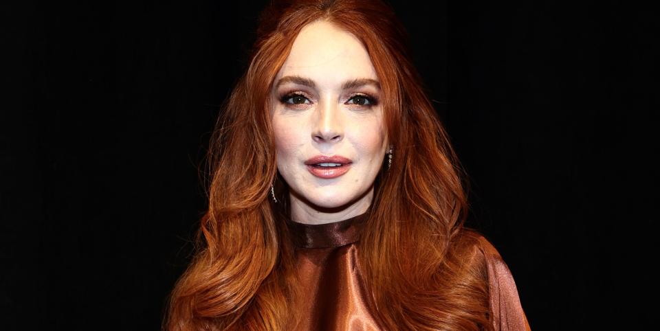 lindsay lohan, a woman stands looking at the camera, long red hair worn down in loose waves, wearing a metallic brown top, cape and trouser suit