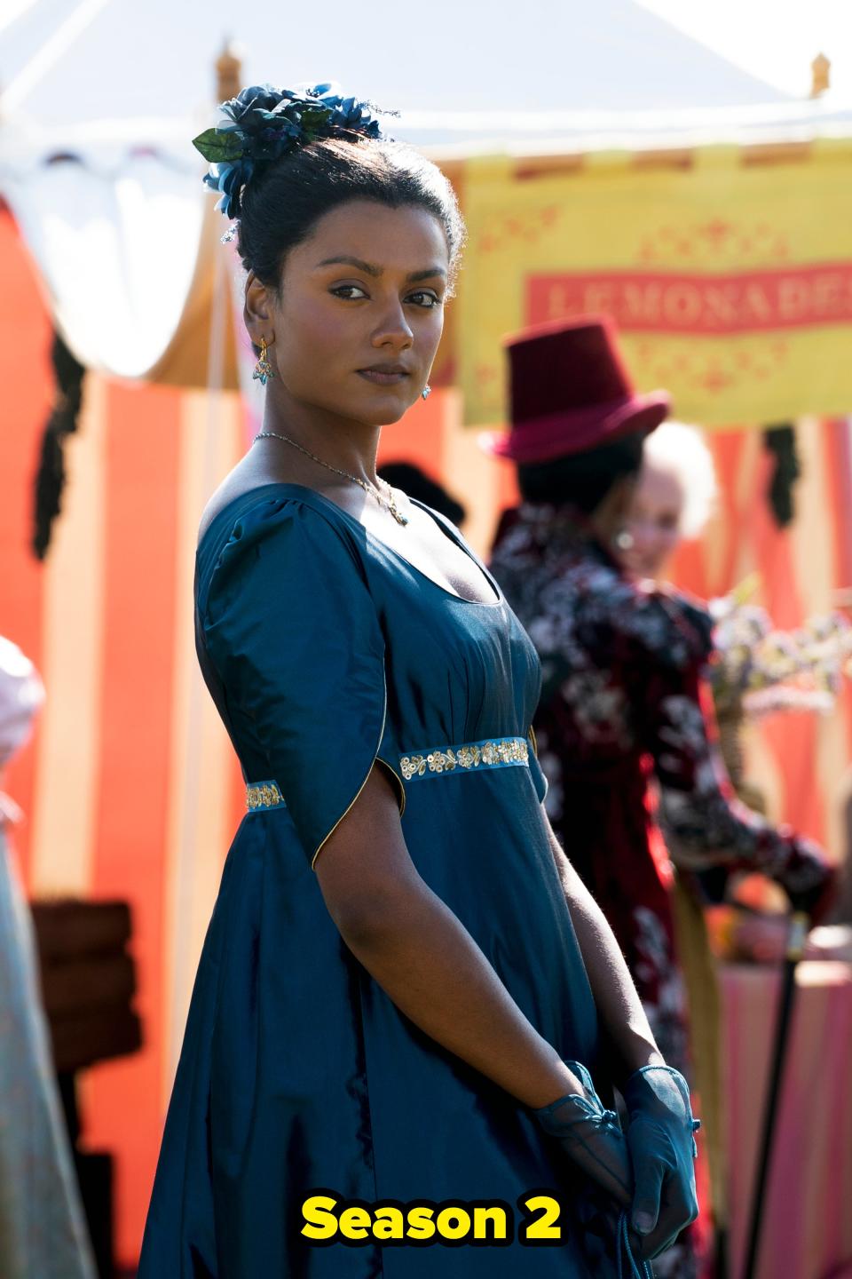 Kate in a period dress scene from Bridgerton, wearing an empire waist gown with cap sleeves and gloves, an updo hairstyle, and floral hair accessory. Background extras included