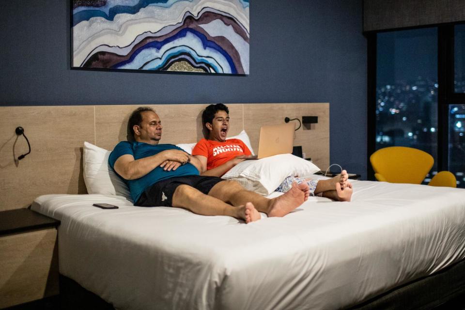 A boy yawns while he and his dad sit on a bed in a hotel.