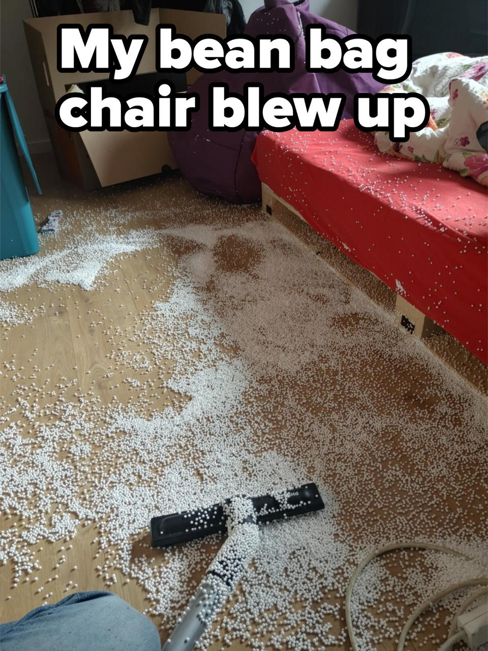 A vacuum cleaner is used to clean up a large amount of white foam beads scattered across a bedroom floor, with a messy bed and boxes in the background