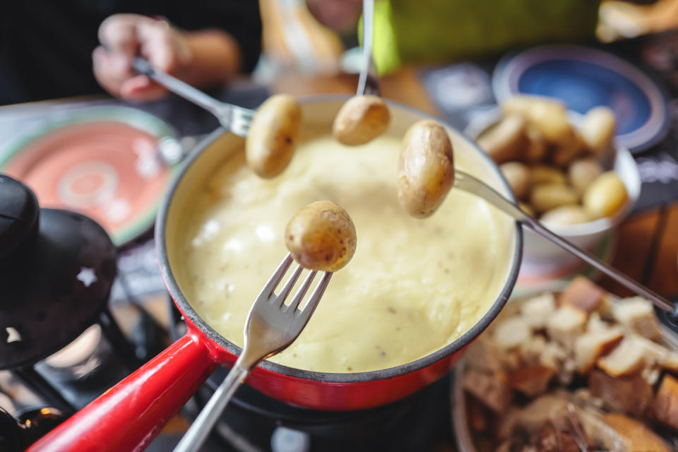 A person dipping a potato on a fork into a fondue pot surrounded by plates of food
