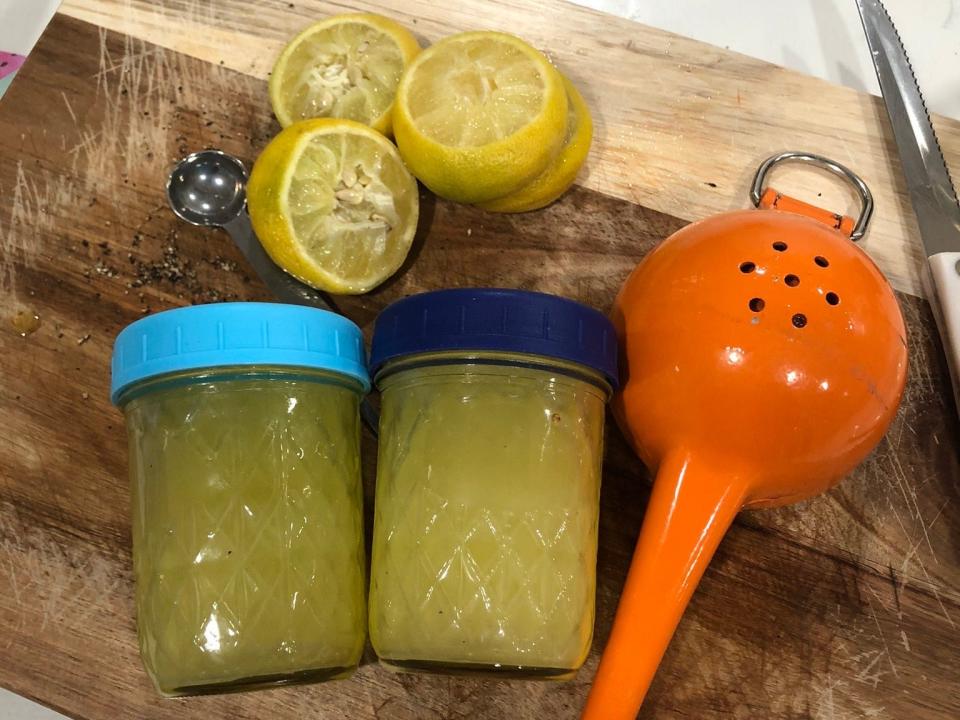 Two jars of lemon vinaigrette, slices of lemons, a measuring spoon, a knife, and a lemon squeezer on a wood cutting board.