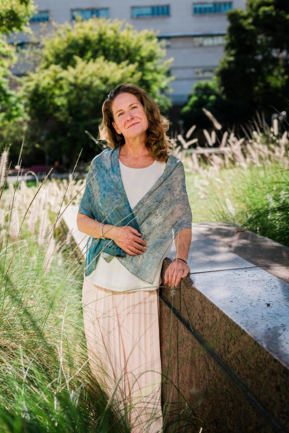 A woman wearing a teal shawl standing amid grasses and other plants, with one hand on a low marble wall