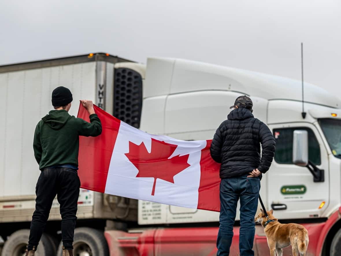 Two men outside Kamloops B.C. hold a Canadian flag Saturday as a semi-trailer passes by. A convoy of trucks is heading to Ottawa to protest a federal vaccine mandate for cross-border travel for truckers. (Candice Camille  - image credit)