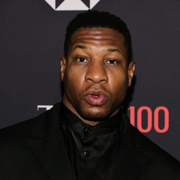 Jonathan Majors in a black suit and shirt at an event, posing for the camera against a backdrop with sponsor logos