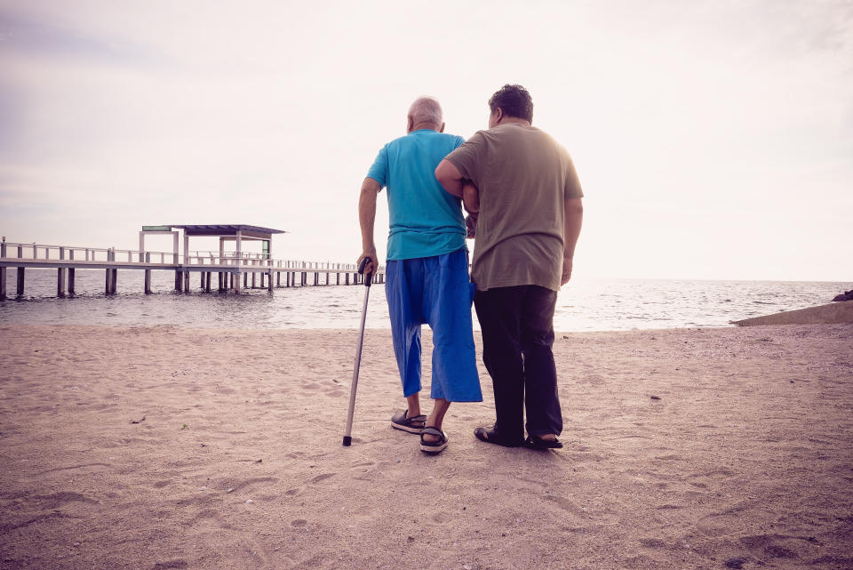 On a beach, with a pier in the background, a man helps support an elderly man walking with a cane.