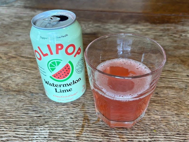 An open can of watermelon-lime Olipop next to a small, clear glass with pink liquid inside. Both are sitting on a wooden table.