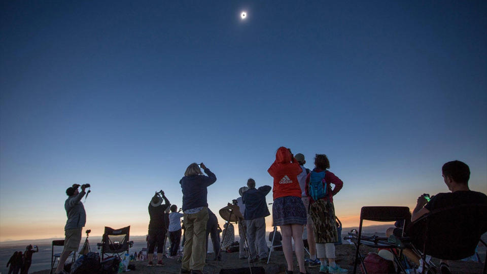 a crowd of people in a field look up at a solar eclipse taking place in a clear sky above