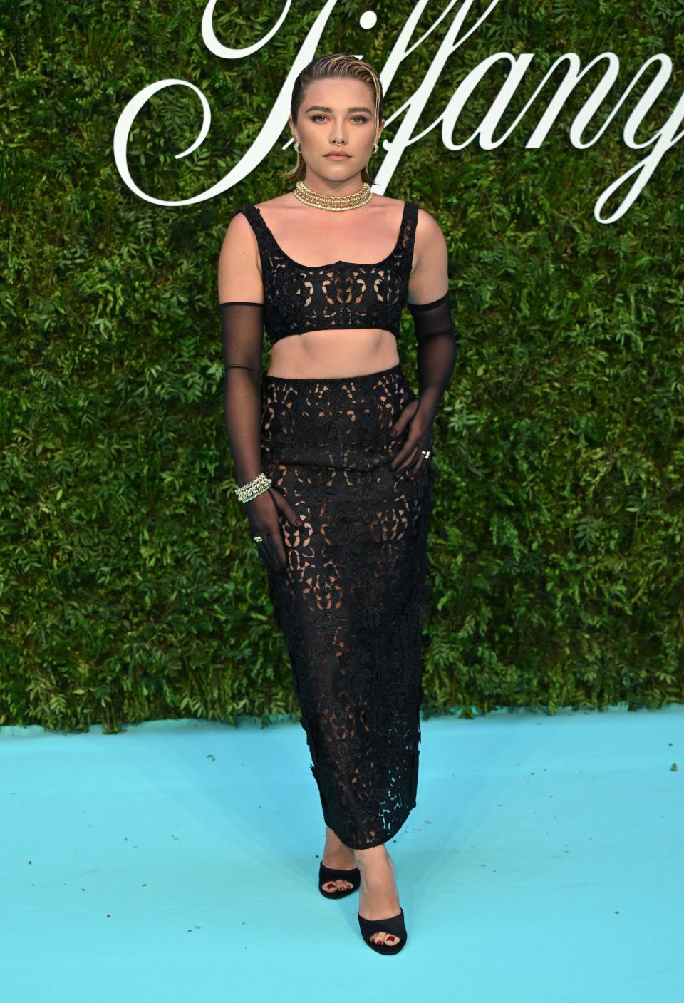 Florence Pugh stands on a blue carpet in front of a greenery wall in a black two-piece.