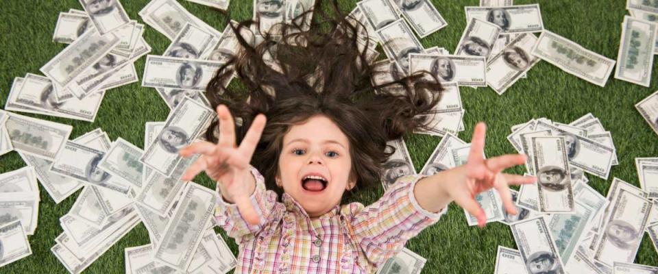 Top view photo of beautiful little girl lying on green grass and dollars. Girl reaching hands to camera