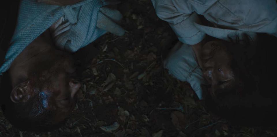 danny (left) and amy (right) facing each other while lying on the ground in beef. they're both wearing white clothes — danny draped in a blanket or cardigan, and amy in a canvas-like coat — as they lay together on the forest floor at night. both of them have dried blood on their faces