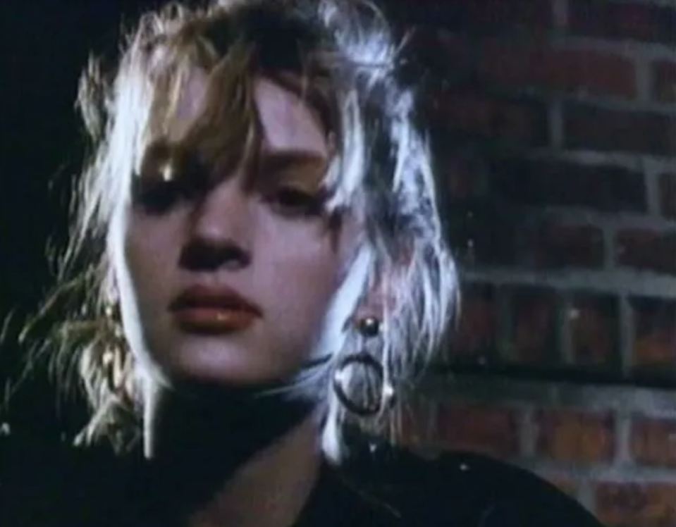 A screenshot of a very young Uma Thurman, taken from a low angle at night in an outdoor alleyway