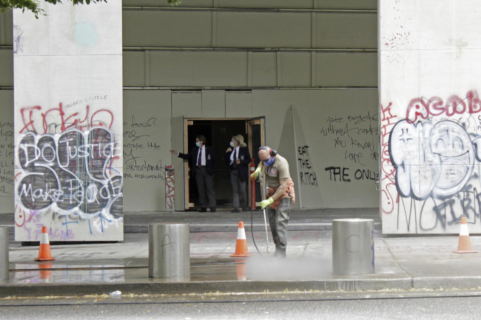 FILE - In this July 8, 2020, file photo, a worker washes graffiti off the sidewalk in front of the Mark O. Hatfield Federal Courthouse in downtown Portland, Ore., as two agents with the U.S. Marshals Service emerge from the boarded-up main entrance to examine the damage. Oregon's largest city is in crisis as violent protests have wracked downtown for weeks. (AP Photo/Gillian Flaccus, File)