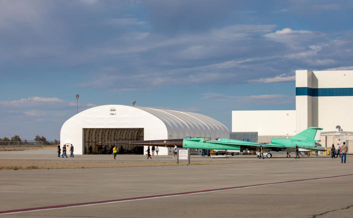 Profile of a plane with pale green undertail coverts with smooth, sharp edges and corners and a long, pointed black nose, which is rolled out of the hangar.  People are scattered around the runway.