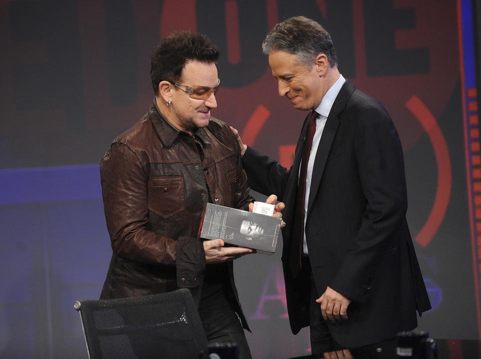 FILE- This Wednesday, Nov. 30, 2011 file photo shows U2 frontman Bono, left, as he presents a pair of headphones to host Jon Stewart during a taping of "The Daily Show with Jon Stewart" in New York. When iPods hit the scene 10 years ago, the small, white ear buds that came with the devices became the symbol for listening to music on the go. Today, that's changing. (AP Photo/Brad Barket, File)