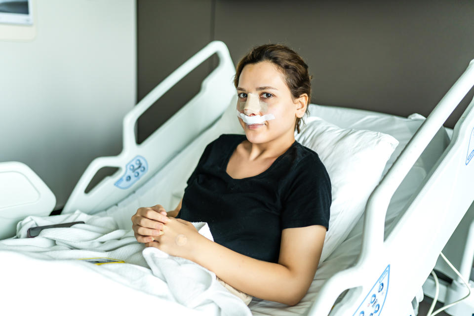 A person with a medical bandage on their nose rests in a hospital bed, looking at the camera. Unknown name