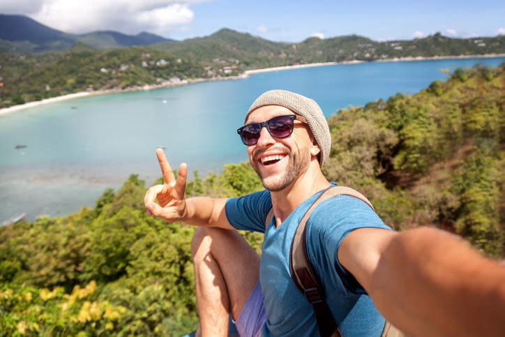 Smiling man sitting near a large body of water and taking a selfie while giving the peace sign